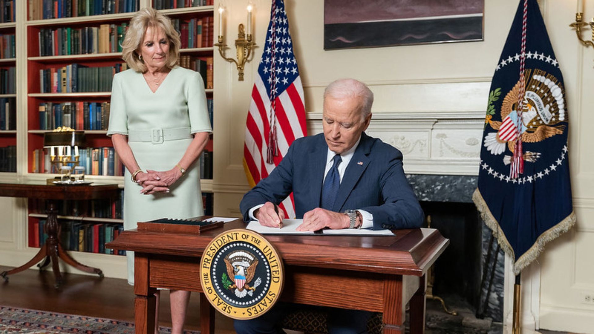 Joe Biden signs a bill at a desk with Dr. Jill Biden standing nearby