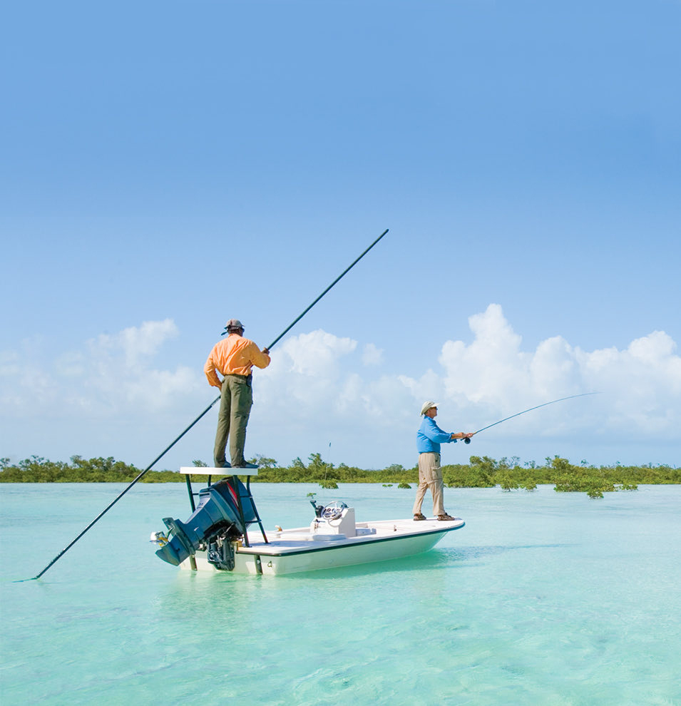Two men stand on a small fishing boat in shallow, clear blue waters. Water reeds line the horizon behind them, beneath and almost cloudless blue sky