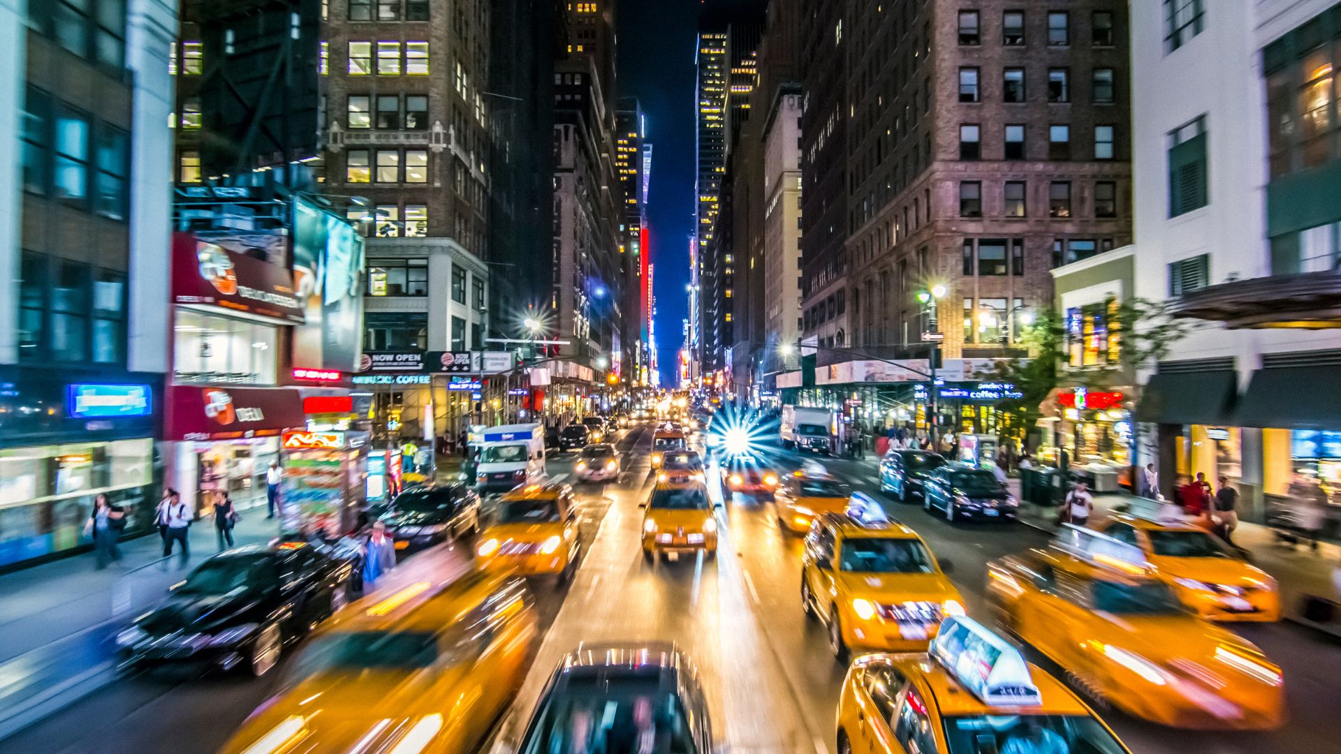 A busy city street with cars moving so fast they are blurred but the street has so many cars and it looks like New York so there's no way traffic isn't at like a complete standstill so that effect is probably misleading. Also, lots of taxis on the road.