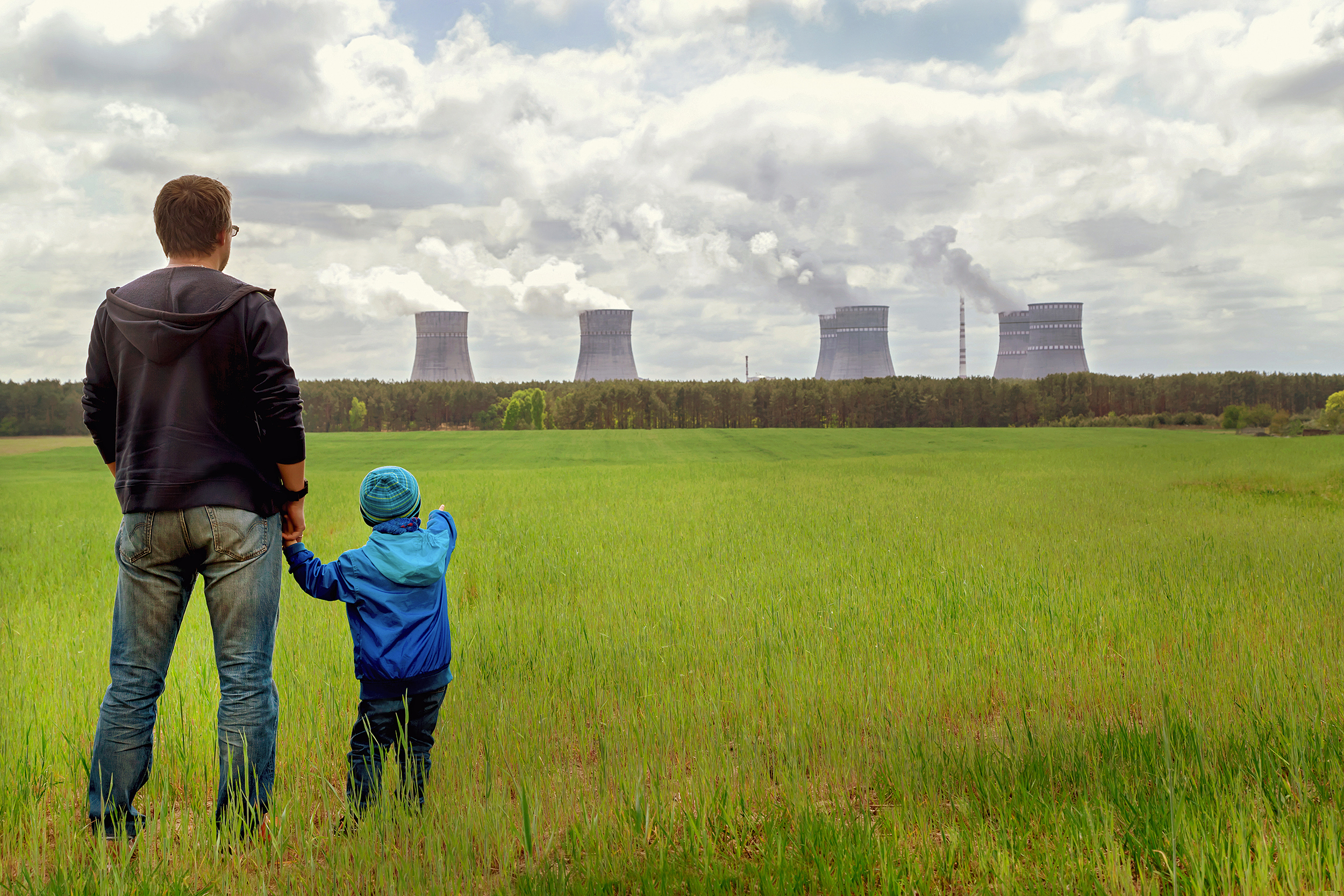 A parent and child stand in a green field looking at billowing smoke stacks in the background