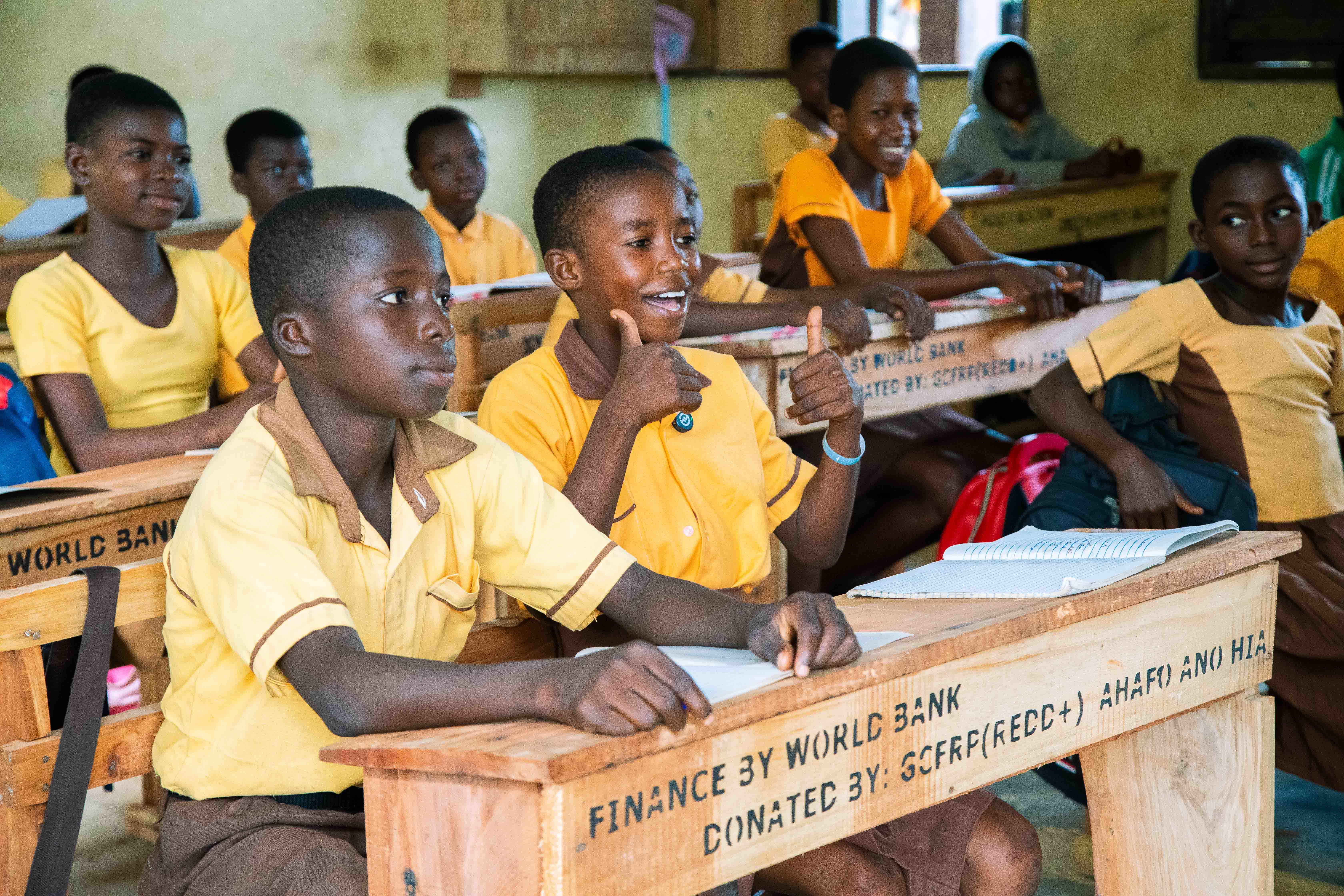 Children sitting at desks from the World Bank. One is smiling and giving a double thumbs up.