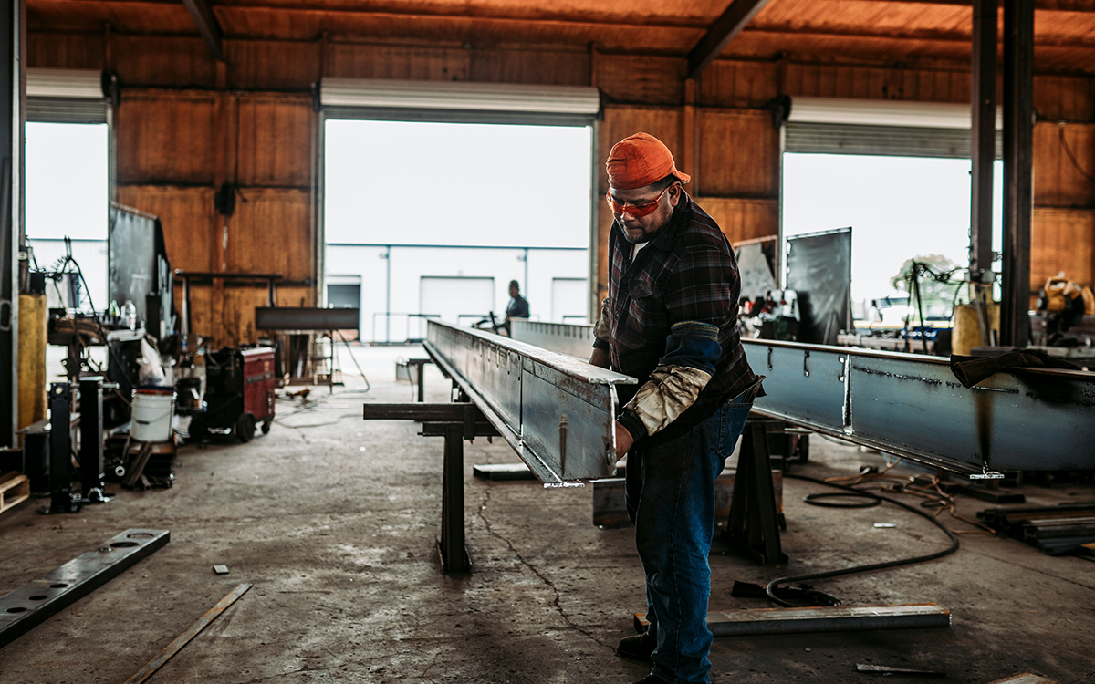 A worker in a metal fabrication shop positions a long steel I-beam on support stands