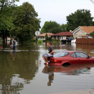 A man sitting on top of car in the middle of flooded area.