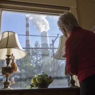 A women looks out of her living room window at a power plant billowing smoke into the air