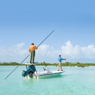 Two men stand on a small fishing boat in shallow, clear blue waters. Water reeds line the horizon behind them, beneath and almost cloudless blue sky