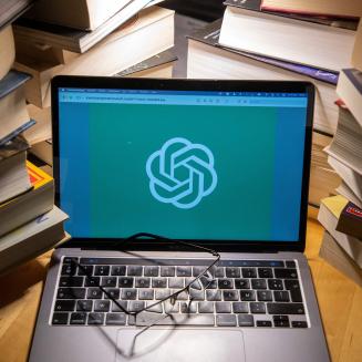 A laptop surrounded by books with the ChatGPT logo on the screen and a pair of glasses on the keyboard