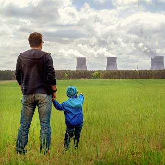 A parent and child stand in a green field looking at billowing smoke stacks in the background