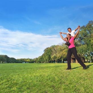 A woman with a child on her shoulders, both smiling, in a grassy field on a sunny day.