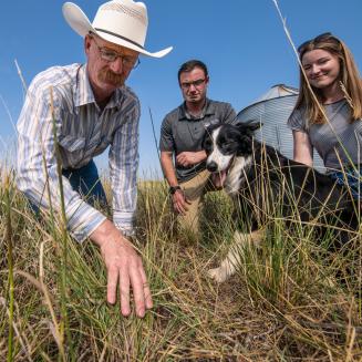 Two people and a dog look on as a person in a cowboy hat looks at the soil