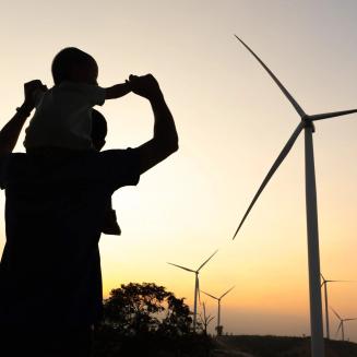 Person holding baby on their shoulders silhouetted against a yellow sky with windmills in the background