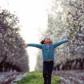 Excited 5 years old little girl walking with wide open raised arms among of almond trees field