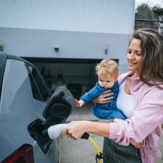 Caregiver holds a baby while charging an electric car in a driveway.