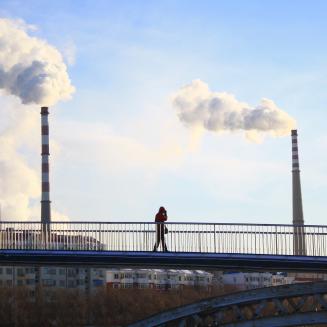 People walk across a bridge as industrial smokestacks release smoke into a blue sky.