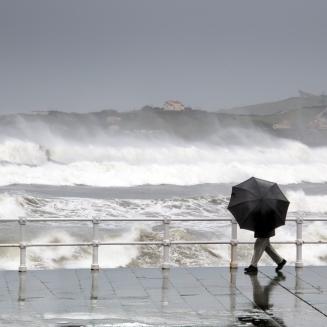 Person with umbrella walking on wet boardwalk in front of a raging ocean