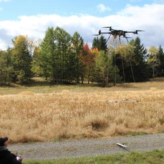 A small team of people operates a drone outdoors, in an area with fields and woods.