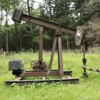 Oil and gas infrastructure in a grassy area, with woods in the background.