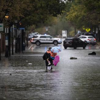 A man carries two children holding colorful umbrellas across a flooded city street.