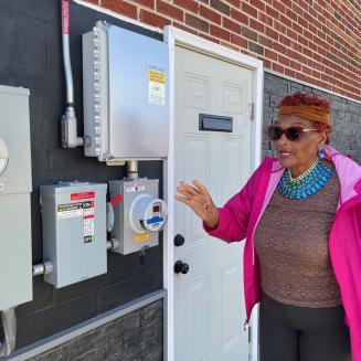 Mary West standing outside of her house pointing to the electric meter.