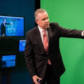 John Morales standing in front of a greenscreen doing the weather.