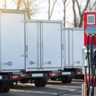 A red electric vehicle charging station stands in the foreground with multiple black charging cables and connectors, while several white box trucks are parked in a row behind it outdoors.