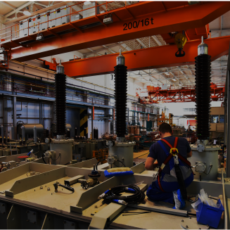 Interior of a large industrial factory filled with heavy electrical equipment. In the foreground, a worker wearing a safety harness kneels on a metal platform, installing or inspecting components such as cables and tools