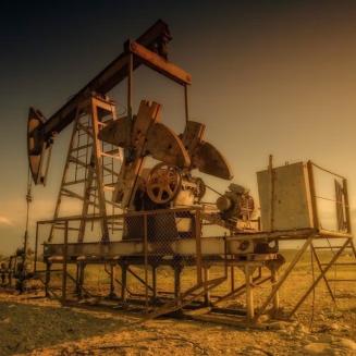 A large oil pumpjack stands in a dry, open field under warm golden light, likely near sunset. The metal structure is elevated on a platform with a ladder, fencing, exposed machinery, and pipes connected to the wellhead below. The ground is rocky and barren, with sparse vegetation. Additional pumpjacks are visible in the distance against the flat horizon. The image has a sepia-toned color palette, highlighting the industrial equipment and expansive landscape.