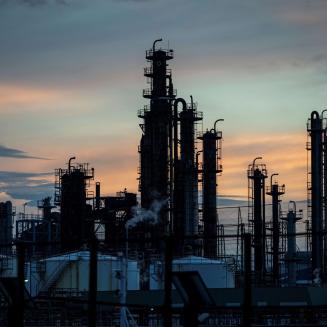 An industrial oil refinery or petrochemical processing facility is silhouetted against a sunset sky. Multiple tall distillation towers, storage tanks, pipes, and metal structures rise above the complex, with faint steam visible from some equipment. The sky is filled with soft blue, gray, and orange hues, creating a contrast with the dark outlines of the machinery. Power lines and additional industrial infrastructure are visible throughout the scene, emphasizing the scale of the facility.