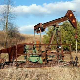 A weathered oil pumpjack with visible rust and peeling paint stands in a dry grassy field. Bare deciduous trees and evergreen shrubs surround the site, with rolling farmland and a partly cloudy blue sky in the background. The aging equipment suggests a low-producing or inactive oil well in a rural setting.