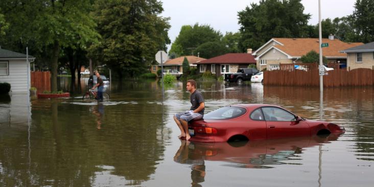 Picture of man sitting on top of car in the middle of flooded area.