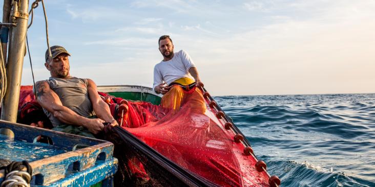 Two fishers on a boat pull fishing gear through open water.