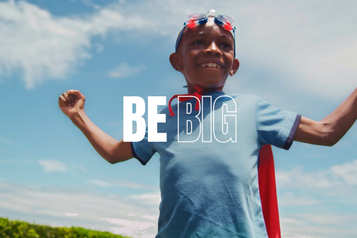 "BE BIG" text on photo of boy with superhero cape with arms raised and blue sky