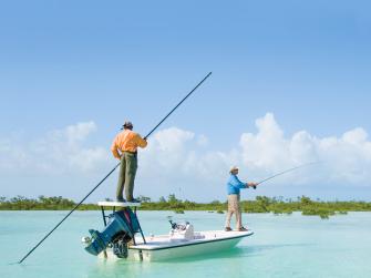 Two men stand on a small fishing boat in shallow, clear blue waters. Water reeds line the horizon behind them, beneath and almost cloudless blue sky