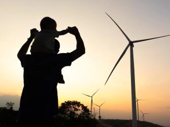 Person holding baby on their shoulders silhouetted against a yellow sky with windmills in the background