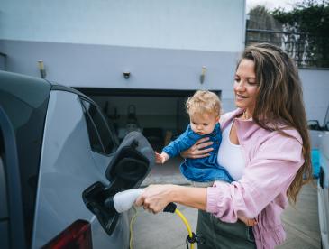 Caregiver holds a baby while charging an electric car in a driveway.