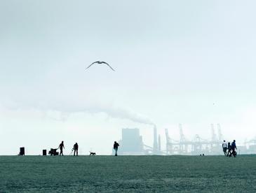 People in a field, with a factory in the background releasing smoke.