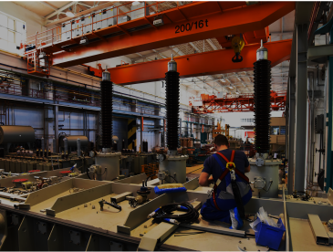 Interior of a large industrial factory filled with heavy electrical equipment. In the foreground, a worker wearing a safety harness kneels on a metal platform, installing or inspecting components such as cables and tools