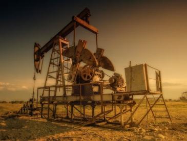 A large oil pumpjack stands in a dry, open field under warm golden light, likely near sunset. The metal structure is elevated on a platform with a ladder, fencing, exposed machinery, and pipes connected to the wellhead below. The ground is rocky and barren, with sparse vegetation. Additional pumpjacks are visible in the distance against the flat horizon. The image has a sepia-toned color palette, highlighting the industrial equipment and expansive landscape.