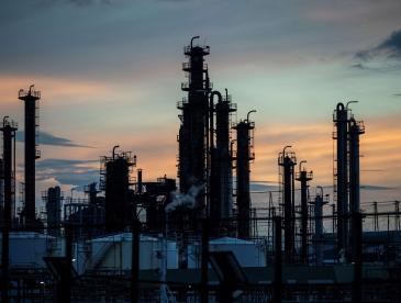 An industrial oil refinery or petrochemical processing facility is silhouetted against a sunset sky. Multiple tall distillation towers, storage tanks, pipes, and metal structures rise above the complex, with faint steam visible from some equipment. The sky is filled with soft blue, gray, and orange hues, creating a contrast with the dark outlines of the machinery. Power lines and additional industrial infrastructure are visible throughout the scene, emphasizing the scale of the facility.
