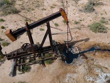 Aerial view of a black-and-orange oil pumpjack standing in a dry, sparsely vegetated landscape. Dark oil-stained runoff extends from the well area across the pale, cracked ground, with pipes and equipment surrounding the pump.