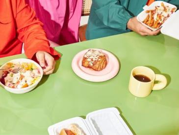 Food and coffee in varying containers and packages, as people sit around the table to eat.