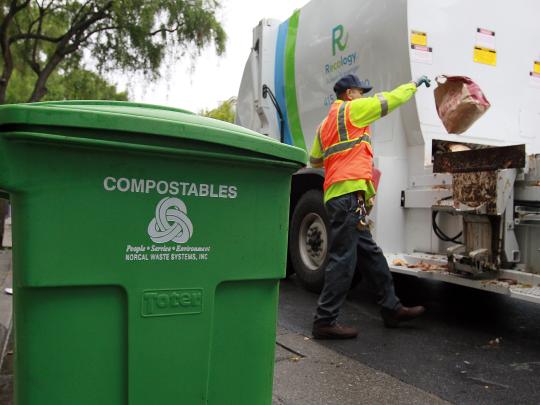 A sanitation worker throws a compost bag into a truck with a green compostables bin in the foreground