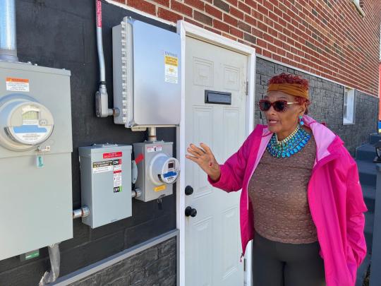 Mary West standing outside of her house pointing to the electric meter.