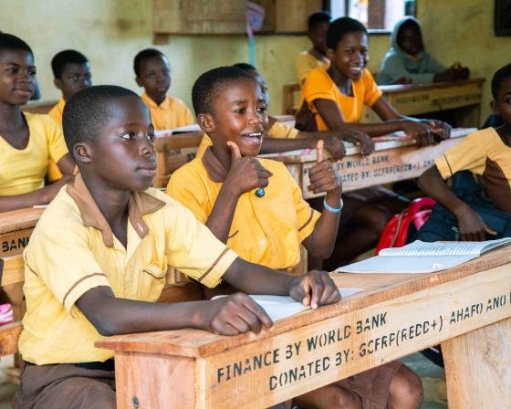 Children sitting at desks from the World Bank. One is smiling and giving a double thumbs up.