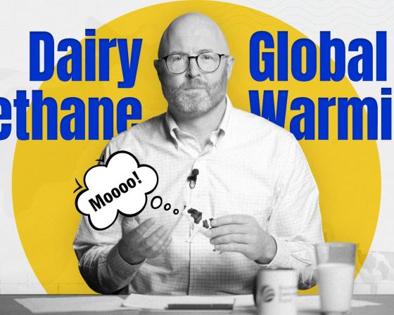 John Tauzel holding a toy cow saying 'moo,' with the words 'dairy methane' and 'global warming' on the screen behind him.