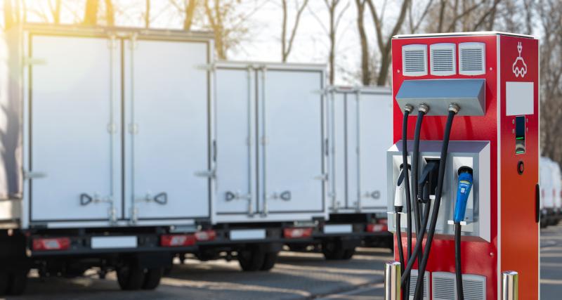 A red electric vehicle charging station stands in the foreground with multiple black charging cables and connectors, while several white box trucks are parked in a row behind it outdoors.