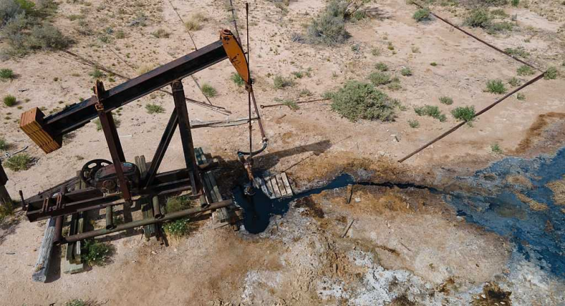 Aerial view of a black-and-orange oil pumpjack standing in a dry, sparsely vegetated landscape. Dark oil-stained runoff extends from the well area across the pale, cracked ground, with pipes and equipment surrounding the pump.